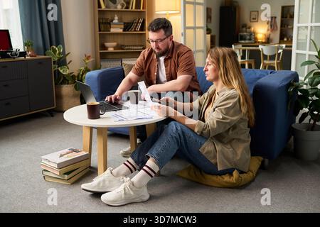 Caucasian young adult man and Caucasian young adult woman sitting on floor in living room reviewing tax documents together Stock Photo