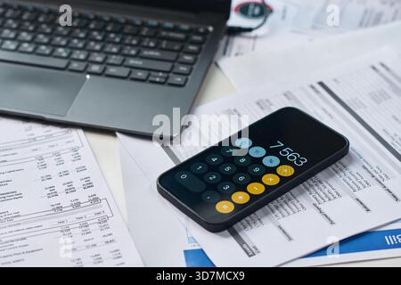 Smartphone displaying calculator app lying on financial documents near laptop, calculating tax payments and analyzing expenses during tax season, close up of modern workspace Stock Photo