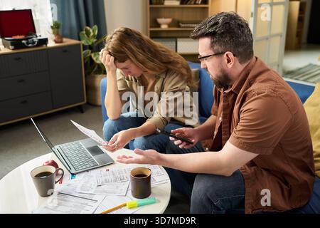 Caucasian young woman and Caucasian young man sitting at table reviewing tax documents and using laptop, both appearing stressed while discussing financial paperwork and deadlines Stock Photo