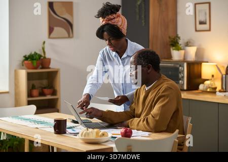 Middle aged Black man sitting at table using laptop and calculator while young Black woman standing beside him holding paper, both reviewing financial documents and paying taxes together Stock Photo