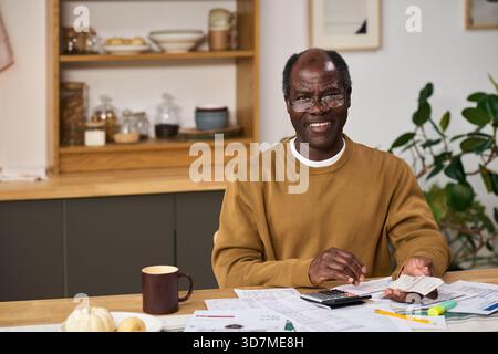 Middle aged Black man sitting at table reviewing financial documents and using calculator while preparing taxes at home, smiling and looking slightly off camera, surrounded by paperwork Stock Photo