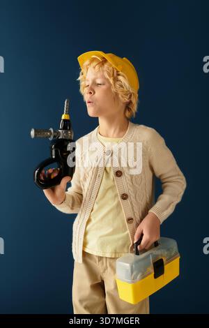 Blonde boy wears a yellow hard hat, pretending to use tools in a playful studio environment. Stock Photo