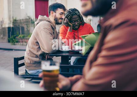 Young multi-ethnic friends sitting at an outdoor cafe table chatting and smiling while drinking coffee. Stock Photo
