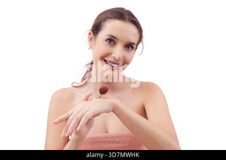 Smiling young woman tapping a makeup brush on the back of her hand before applying blush while looking into the camera. Clean, minimal beauty routine Stock Photo