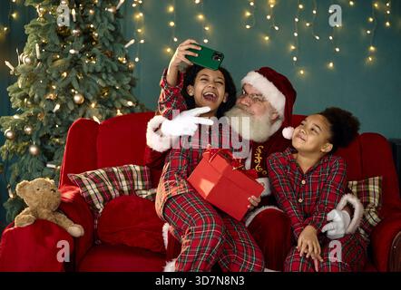 santa claus and two african american sisters taking selfie on red couch at home Stock Photo