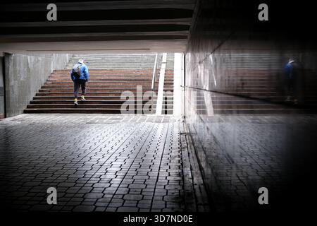 A person in a blue jacket walks up wet stairs in an urban tunnel, reflecting on the walls. Stock Photo
