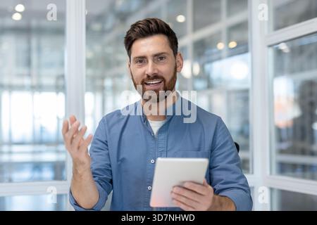 Smiling businessman in smart-casual attire holding a tablet and gesturing while presenting ideas during a virtual meeting or webinar in a modern office setting Stock Photo