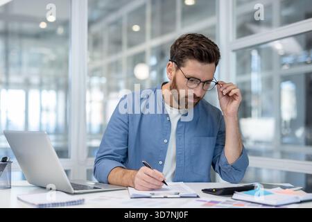 Serious male professional analyzing financial data, calculating expenses, and taking notes on paperwork, demonstrating dedicated work ethic and attention to detail in a modern office setup Stock Photo