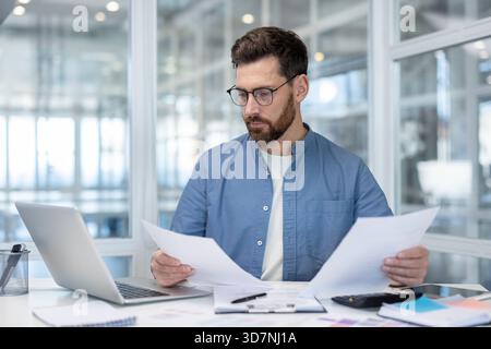 Young businessman wearing glasses carefully reviewing financial paperwork and working with laptop at desk, managing important financial data and administrative tasks in an office environment Stock Photo