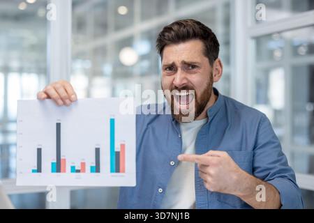 Stressed businessman in modern office angrily pointing at a bar chart with declining results, shouting in frustration over negative financial data and report failure Stock Photo