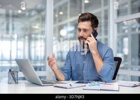 Young bearded businessman communicating on smartphone, multi-tasking and managing business tasks remotely while sitting at a desk with a laptop and documents in a contemporary office Stock Photo