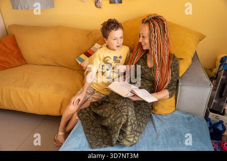 A happy mother with colorful dreadlocks smiles at her excited son as they sit together on a yellow sofa, sharing a book and enjoying a joyful moment o Stock Photo