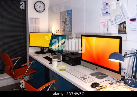 Modern shared office workspace with multiple computer monitors, apple macs, wall clock, documents, and ergonomic chairs in a compact organized setting Stock Photo