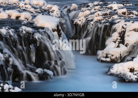 View of cascading waterfalls plunge into an icy riverbed framed by snow-laden rocks, creating a serene winter landscape, Hlauptunga, Blaskogabyggd, Ic Stock Photo