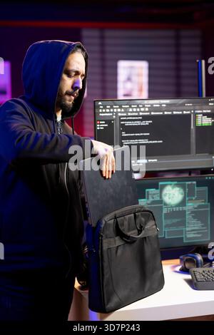 Hacker in hideout putting laptop in backpack after stealing sensitive information using VPN on dark web. Man packing notebook after finishing breaking into systems to take restricted data Stock Photo
