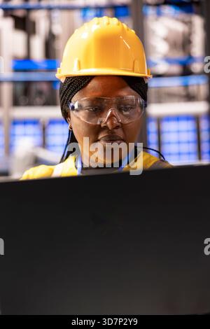 Closeup of african american engineer using personal computer in modern facility. Selective focus on black woman wearing safety goggles and industrial helmet, looking at her laptop in a factory. Stock Photo