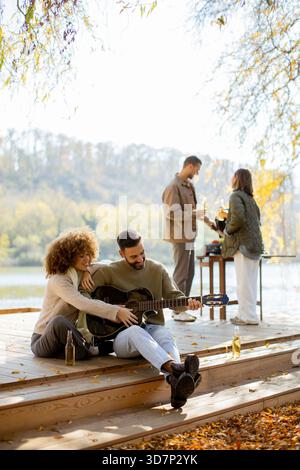 Two friends enjoy music on the wooden dock while others prepare food nearby, surrounded by colorful autumn leaves. Stock Photo