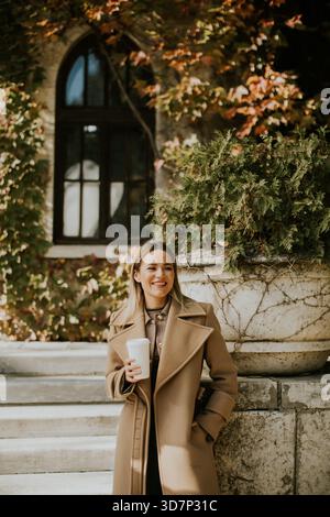 A young woman stands on steps with a big smile while holding a warm drink. She wears a fashionable coat, and colorful leaves create a vibrant autumn b Stock Photo