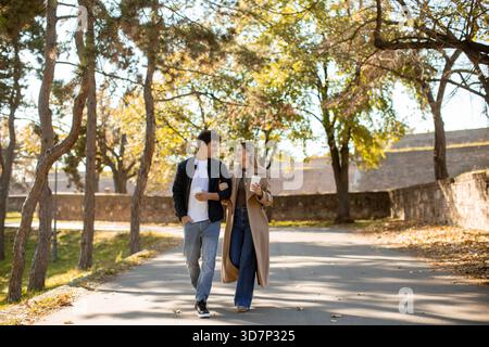 A young couple walks hand in hand in a park surrounded by trees with autumn leaves. They are engaged in a pleasant conversation while sipping coffee, Stock Photo