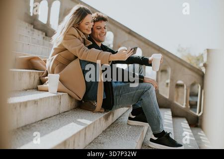 Two people sit closely on stone steps, smiling and looking at a phone together. They enjoy a sunny day, with one holding a cup and the other relaxed i Stock Photo