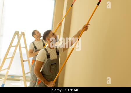 Two men are painting a light-colored wall in a spacious room with large windows. They use long brushes and stand on a ladder, concentrating on their t Stock Photo