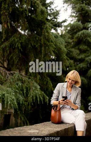 A woman in light clothing sits on a stone wall surrounded by tall trees. She is focused on her smartphone, enjoying a peaceful moment in nature. Stock Photo