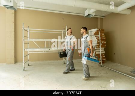 Two construction workers are walking through a newly renovated room. They carry toolboxes and discuss the ongoing project. The space is empty, with sc Stock Photo
