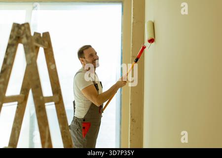 A man is smiling while using a paint roller to apply a fresh coat of paint to a wall. A ladder is seen nearby, and sunlight floods the room, creating Stock Photo