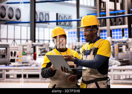 African american engineers at smart manufacturing plant analyzing automated production systems using laptop. Male and female technicians improving digital automation assembly line performance. Stock Photo