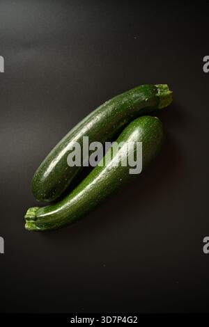 Top-down shot of two glossy green zucchinis placed diagonally and overlapping on a deep black background. Ideal for themes of healthy eating, veganism Stock Photo