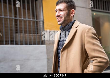 A cheerful man strolls down a lively street, dressed in a warm coat. Sunlight highlights the modern buildings around him. He appears relaxed, enjoying Stock Photo