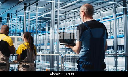 Smart factory expert using laptop software to test automated systems and ensure reliability. Worker uses program on notebook to identify bottlenecks and optimize gear based on sensors data, camera A Stock Photo