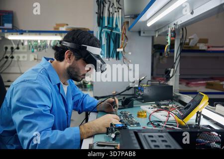 A man soldering technician in a blue lab coat works on electronics and devices Stock Photo
