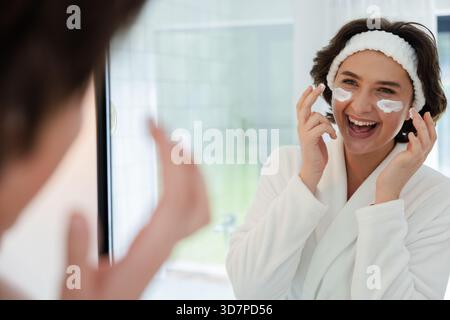 Woman wearing bathrobe applying skincare cream under eyes at home bathroom mirror, copy space Stock Photo