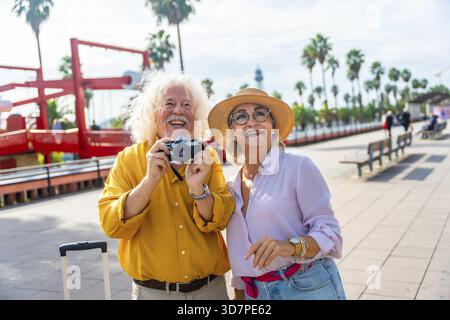 Happy senior couple enjoying their vacation and exploring a sunny city while traveling, capturing memories with a vintage camera on a beautiful boardw Stock Photo