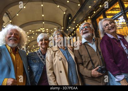 Group of happy senior friends smiling while walking together in a city street at night, experiencing urban life and travel with string lights overhead Stock Photo