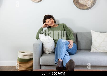 Woman leaning on white throw pillow, resting head on hand on gray sofa in living room Stock Photo