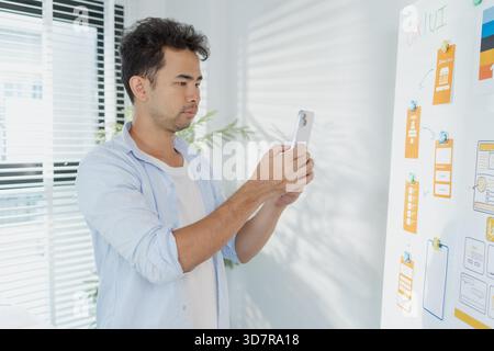 A creative designer works on UX/UI planning, analyzing sketches and prototypes on a whiteboard while using a laptop. The process shows design thinking Stock Photo