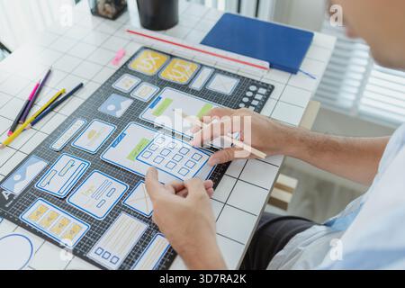 A designer is developing UX UI layouts, reviewing mobile app prototypes, and arranging interface elements on paper and board. He is focused on user ex Stock Photo