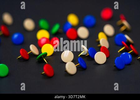 A scattering of colorful thumbtacks (pushpins) in red, blue, green, and beige on a black surface, with a shallow depth of field. Stock Photo