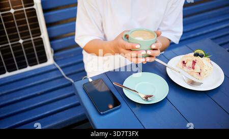 Girl drinking coffee while phone charging from solar panel. Cropped view of woman sitting next to portable solar battery charger. Integration of sustainable renewable energy into everyday life. Stock Photo