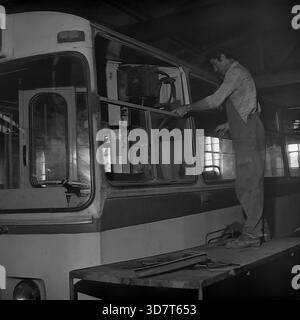 Slavyansk, Ukraine, USSR - 1983: A mechanic wearing denim overalls and a plaid shirt performs bodywork repairs on a Soviet ZiU-9 trolleybus inside the city depot hangar. Standing on a makeshift platform, the worker carefully installs a side window pane into the vehicle's frame. The interior of the workshop is dimly lit with industrial windows in the background. This black and white archival photo captures the manual labor, technical maintenance, and industrial routine of the public transport system in the peaceful Donbas city. Stock Photo