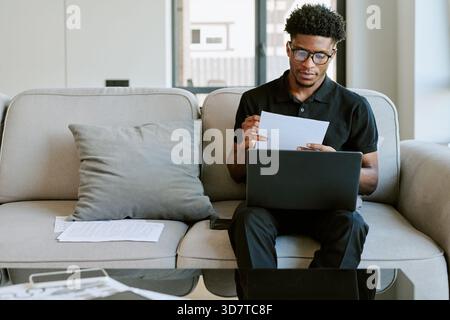 Young Adult Black Man Reading Documents While Working on Laptop in Morning Stock Photo
