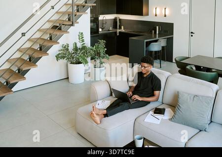 Young Adult Black Man Relaxing on Sofa Using Laptop in Modern Living Room Stock Photo
