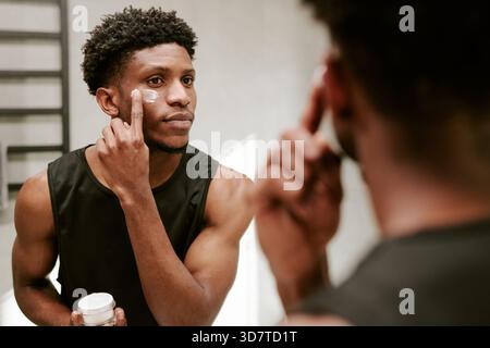 Young Adult Black Man Applying Face Cream While Looking in Bathroom Mirror Stock Photo