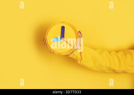A hand in a yellow sleeve holds a modern yellow wall clock against a solid yellow background, symbolizing time and punctuality. Stock Photo