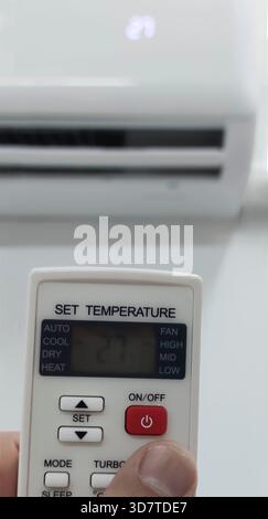 Adjusting conditioner temperature with remote control to 27 celsius degrees. Hand with remote control directed on the wall attached device Stock Photo
