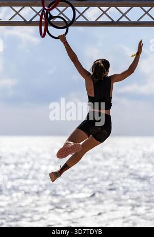 Rear view of a young female athlete balancing as she completes a rings obstacle high above the sea, with a bright sky in the background. Stock Photo