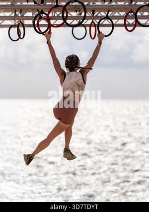 A slender athlete jumps and balances between rings suspended from a scaffold with the crystal-clear, shimmering water of the ocean as a backdrop. Stock Photo