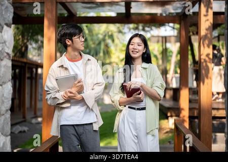 Asian friends or a man and a woman holding books and standing talking together in wooden corridor. Working or Study outdoor, Garden Cafe or Restaurant Stock Photo